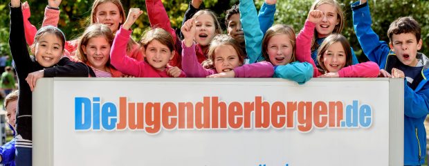 Eine Schulklasse steht hinter dem Schild der Jugendherberge und jubelt