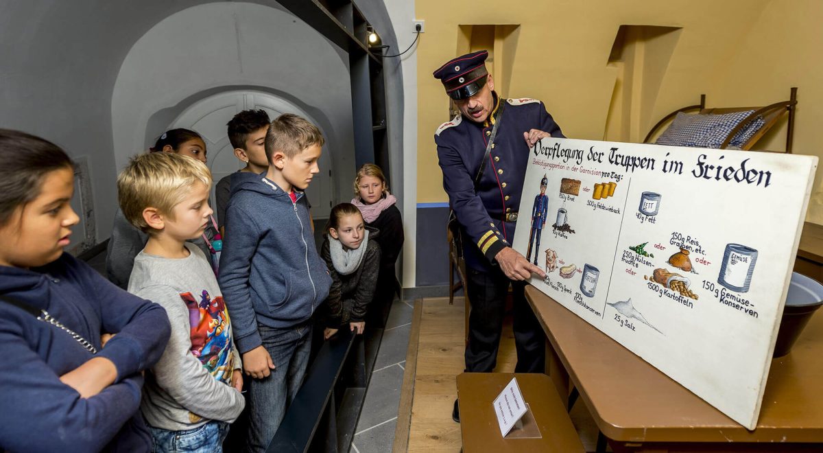 Ein Darsteller in preußischer Uniform erklärt einer Gruppe von Kindern eine Tafel zur Verpflegung der Truppen im Frieden auf Festung Ehrenbreitstein Koblenz.