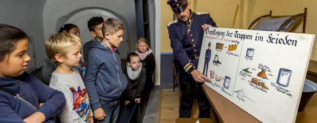 Ein Darsteller in preußischer Uniform erklärt einer Gruppe von Kindern eine Tafel zur Verpflegung der Truppen im Frieden auf Festung Ehrenbreitstein Koblenz.