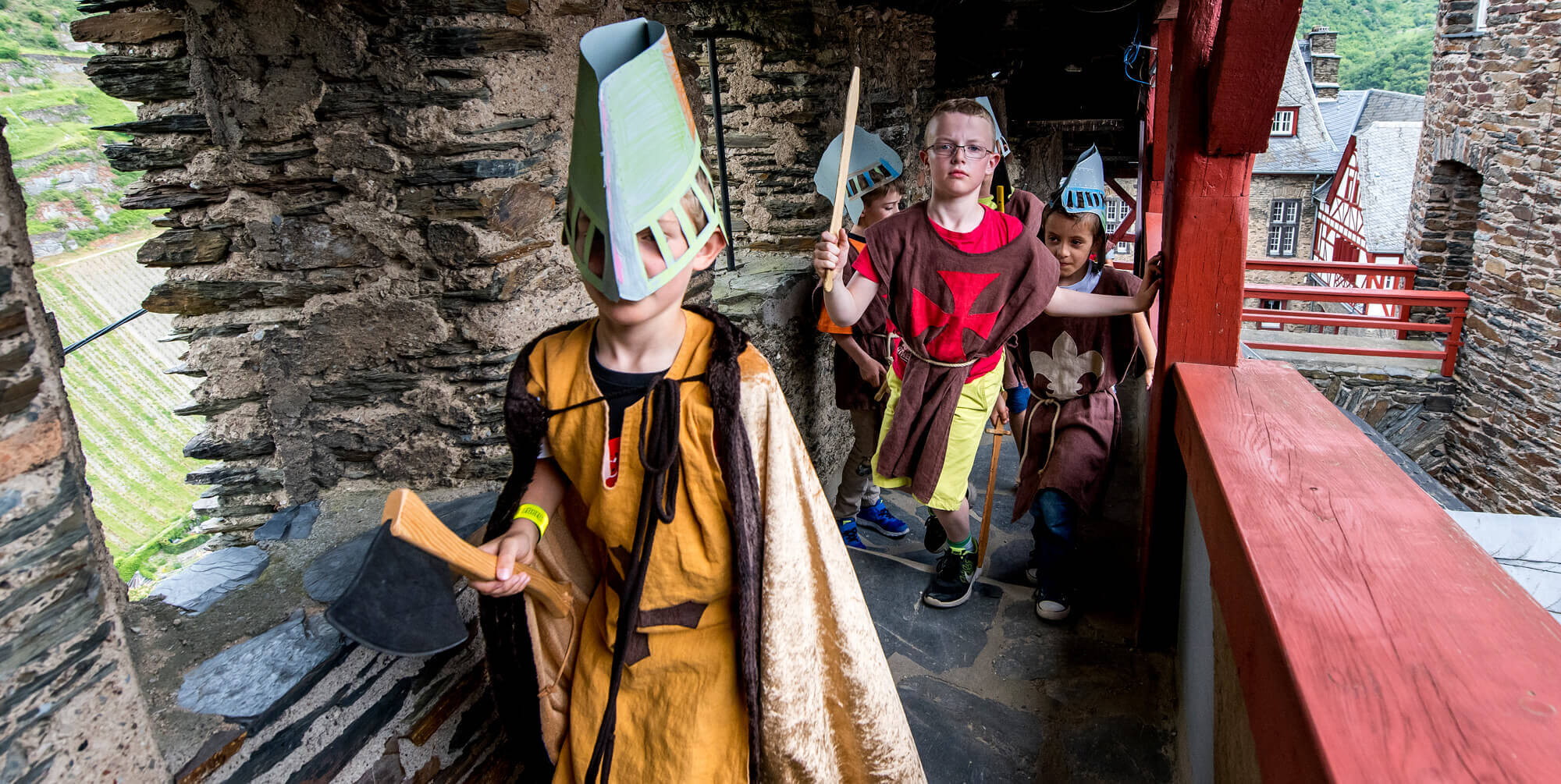 Kinder mit selbstgebastelten Helmen und Holzschwertern erkunden einen schmalen Wehrgang der Burg Stahleck.