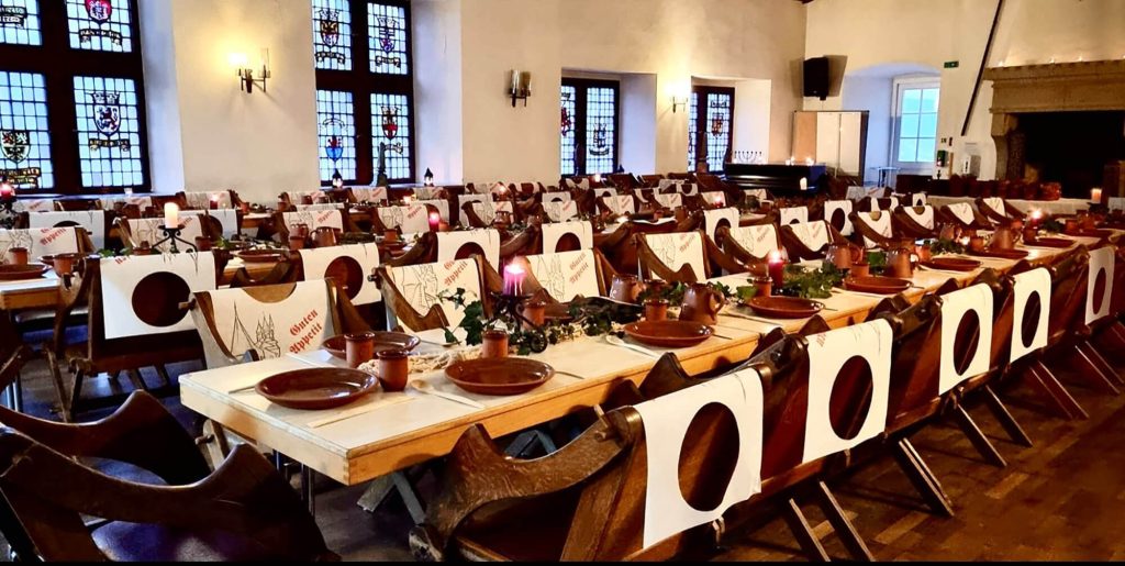 Lange, rustikale Holztische in historischen Saal der Jugendherberge Bacharach mit Wappenfenstern, eingedeckt mit Kerzen und Keramikgeschirr für ein Rittermahl.