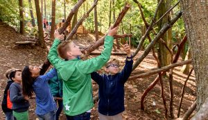 Sechs Kinder sind im Wald und bauen mit langen Ästen eine Hütte.