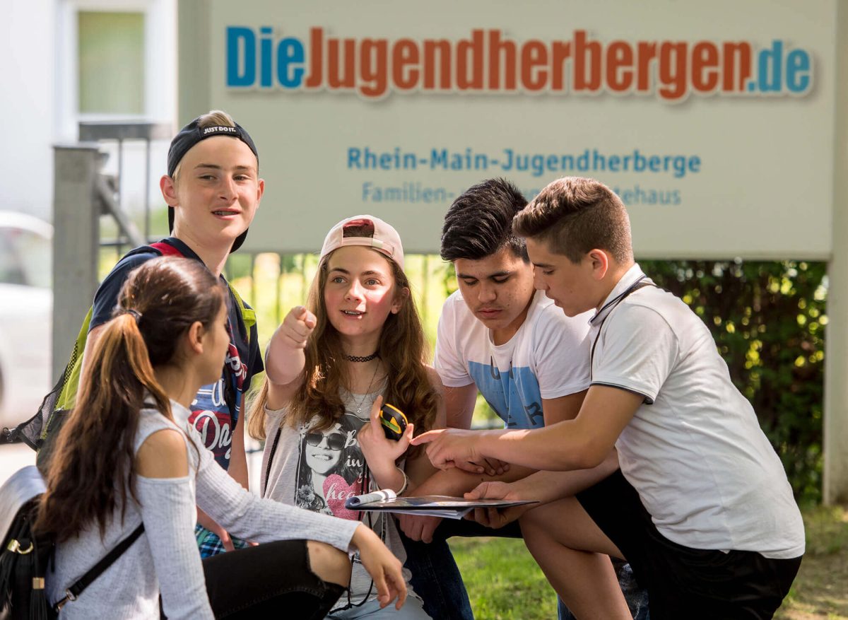 Eine Schülergruppe bespricht den Weg mit Karte und GPS vor dem Schild der Jugendherberge Mainz während eines Klassenfahrtprogramms.