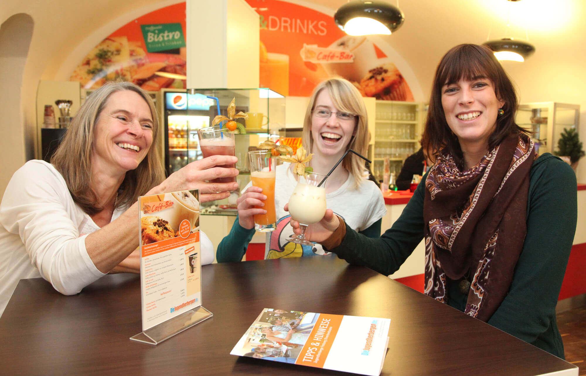 Drei Frauen sitzen fröhlich im Bistro der Jugendherberge Koblenz und stoßen mit bunten Cocktails an.