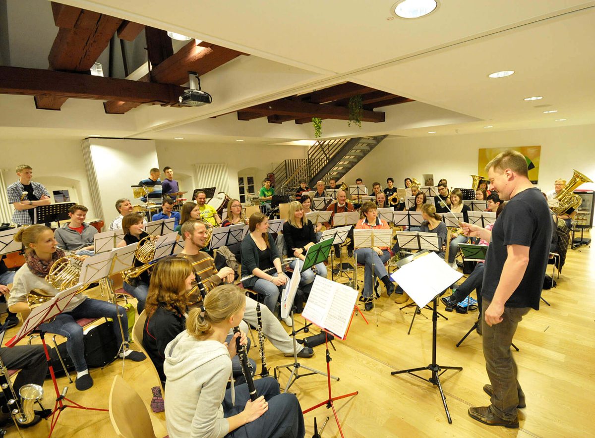 Mit viel Spaß probt eine große Musikgruppe im Aufenthaltsraum der Jugendherberge Kaub. Die Musiker sitzen mit ihren Instrumenten und Notenständer im Halbkreis vor dem Dirigenten.