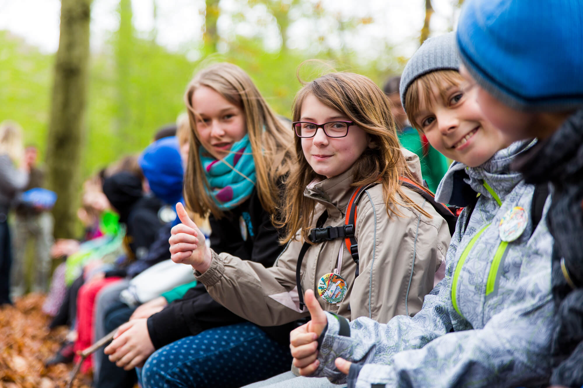 Schüler in Winterkleidung sitzen im Wald und zeigen Daumen hoch